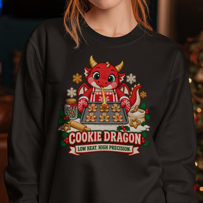 Weihnachtspullover mit einem roten Drachen in gestreifter Schürze und Ofenhandschuhen, der mit einem feinen Flammenstrahl ein Lebkuchenmännchen auf einem Backblech bräunt, darunter der Schriftzug Cookie Dragon.